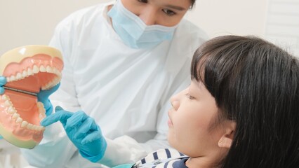 Naklejka premium Asian female pediatric dentist demonstrate toothbrush to little girl with teeth model in dental clinic, well-being hygiene, and professional orthodontic healthcare work in kid hospital.
