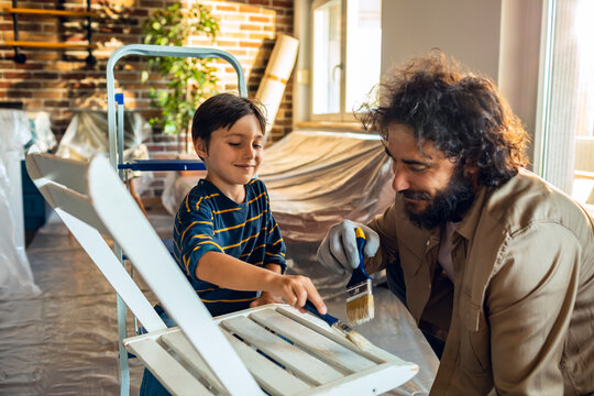 Young caucasian boy and his father painting a wooden chair at home in the living room