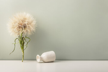 A large dandelion on the table and an overturned small ceramic vase on an olive background.