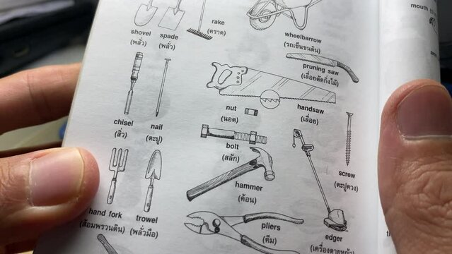 Hand open small handbook for reading with sketch of mechanic and carpentry tools pictures and word translations English - Thai. Study and education concept
