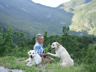 a girl in dreadlocks and two fawn Labradors against the backdrop of mountains. Friendship, walking with dogs