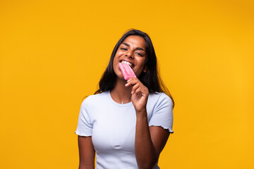 Young Indian woman eating pink popsicle on yellow background. Asian woman eating ice cream. Looking...