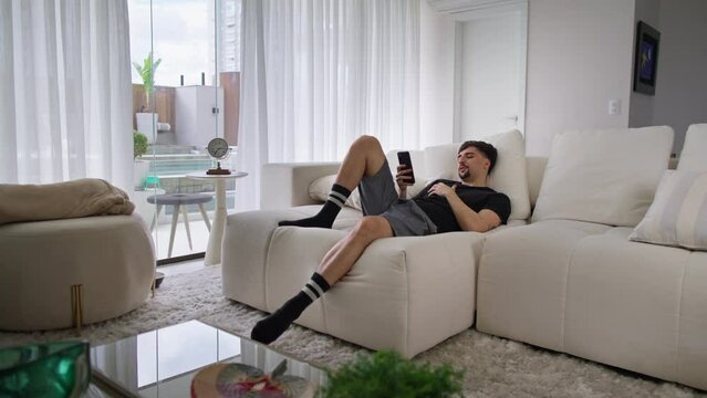 Sleepy young man brownsing on his phone until he falls asleep. Man lying on the couch at home on a lazy morning