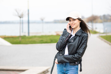 Obraz premium A young pretty girl, dressed in a leather jacket and jeans, spends time alone on the river. The girl holds a mobile phone in her hands and talks