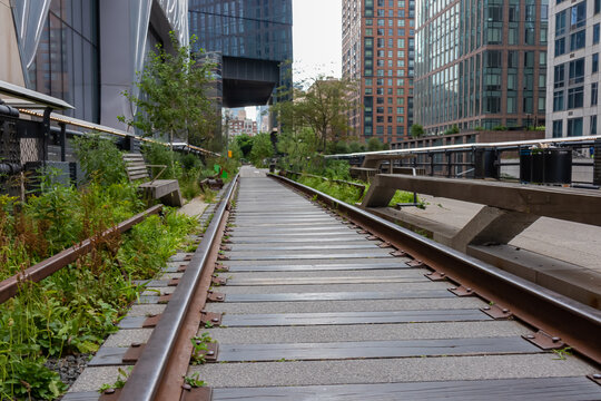 An old railing turned into a pathway - the famous High Line in New York City. The railing is overgrown with greenery. Some benches on the sides. Tall buildings around. Rearranging the cityscape