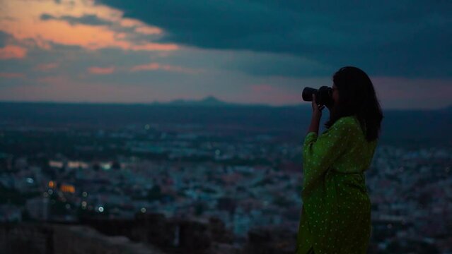 Silhouette shot of young Indian woman taking photos of city with camera while standing on Hill at Bhuj, Kutch, Gujarat. Female photographer shooting during late evening. Travel and holidays concept.