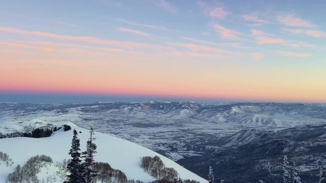 Panning right shot of a stunning winter landscape scene looking down at a cloudy snow covered valley during a golden sunset from the summit of a ski resort in the Rocky Mountains of Utah.