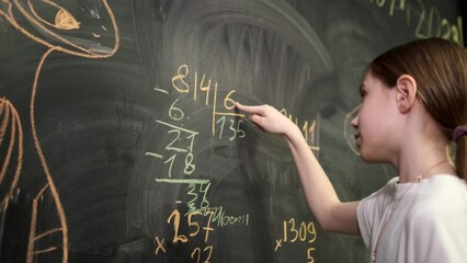 A school-age girl solves mathematical examples at the blackboard during the lesson. Primary school education. Write with chalk on a blackboard.