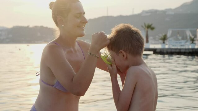 Smiling mother putting on snorkeling mask on her little son before him swimming in sea. Holiday, summer vacation and tourism.