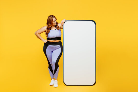 Full body fun young plus size big fat fit woman wear blue top warm up train look at big huge blank screen mobile cell phone isolated on plain yellow background studio home gym. Workout sport concept.