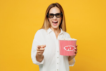 Young laughing fun woman in 3d glasses wear white casual clothes watch movie film hold bucket of popcorn in cinema point index finger camera on you isolated on plain yellow background studio portrait.