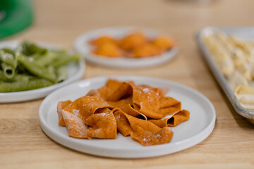 Closeup of handmade pasta, cut into thick, wide, noodles and brightly colored with tomato paste. Other shapes and colors of handmade pasta fill the background.