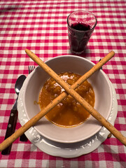 A nice bowl of bean soup, with breadsticks, on a red-checkered tablecloth and a glass of red wine.