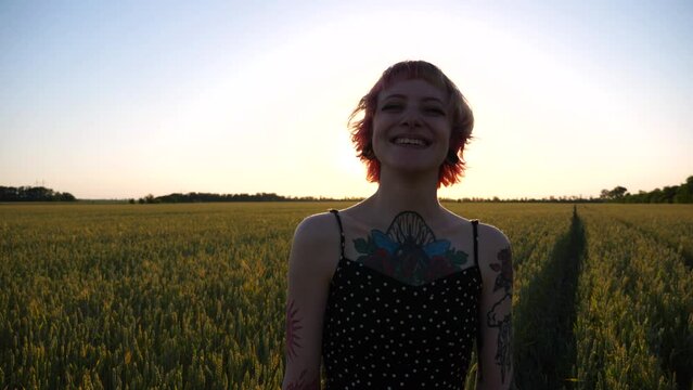 Portrait of smiling hippie woman with ear tunnels with wheat green field at background. Happy punk girl with pink hair looks into camera standing on barley meadow at sunset. Concept of freedom