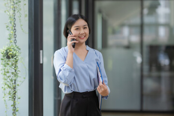 Happy Asian businesswoman talking on mobile phone and holding clipboard in modern office.