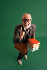 Full length portrait of senior man with beard, teacher wearing vintage suit with big stack of books and brown business bag on green background.