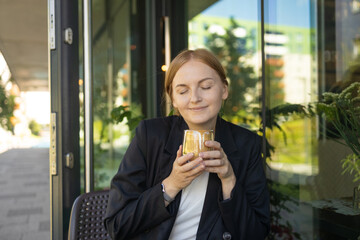 Close up of young female in business casual wear drinking cup of coffee while sitting at small table and enjoying time in street cafe. High quality photo