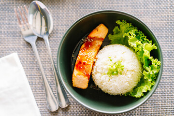 Steamed rice with grilled salmon in a black bowl and spoon on the dining table in a restaurant.