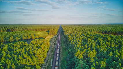 Electric locomotive with railway wagons rides on railroad. Transportation, delivery export and import between cities and countries. Aerial view over train riding through forest.