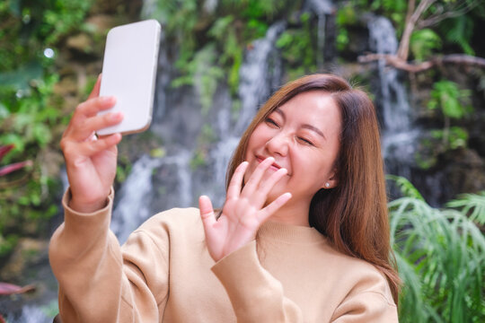 Portrait image of a beautiful young asian woman using mobile phone to take a selfie in the garden with waterfall