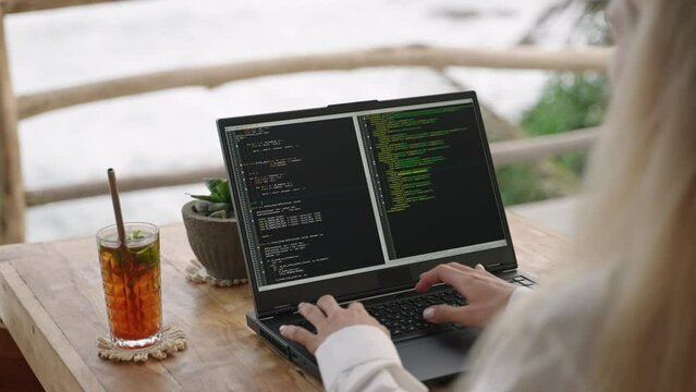 Female soft dev with laptop on remote work day in tropical cafe. Software developer writes program code on computer screen in coworking restaurant with sea view on sunset. Woman coding by ocean.