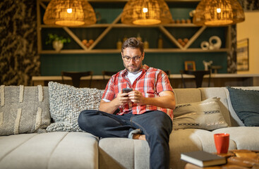 Portrait of man looking at smart phone at living room. Man is talking chatting on the smartphone. Thoughtful man using mobile phone at home. Middle-aged man using phone for web browsing, social media.