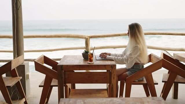 Woman working with laptop in cafe with ocean view, palm trees. Travel blogger typing on computer keyboard sitting in luxury restaurant. SMM manager on remote work day. Female drinks ice coffee by sea.