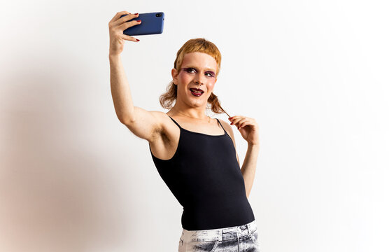 A red-haired non-binary person is posing on a white background in extravagant clothing and takes a picture with the mobile phone while pulling his hair. Concept of social networks. Relate socially.