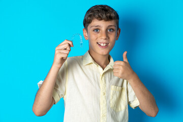 Beautiful kid boy wearing casual shirt holding an invisible braces aligner and rising thumb up,...
