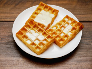 Tasty, golden Belgian waffles covered with sweet condensed milk and served on a white plate on wooden table. Appetizing desert for tea of coffee. Bakery product.