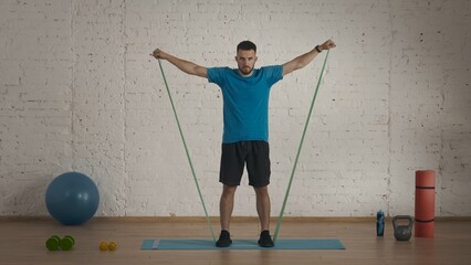 Man fitness trainer in sportswear doing arms up lifting exercise with rubber band for online class at home.