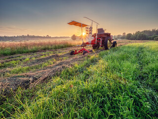 Fototapeta premium Old rusty tractor with grass cutting machine in a agriculture field with tall grass at stunning sunrise. Farming industry. Beautiful nature scene at special time. Nobody.