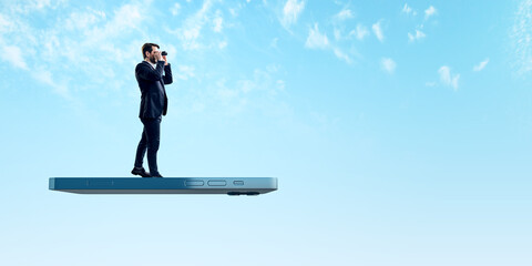 User experiences for mobile concepts. Young businessman with telescope standing on huge flying smartphone on wide bright blue sky background with mock up place.