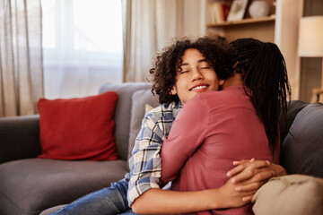 A smiling African teenage boy enjoying a hug with his caring mother, sitting on a couch.