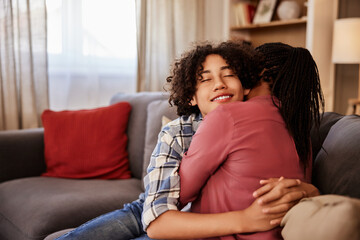 Obraz premium A smiling African teenage boy enjoying a hug with his caring mother, sitting on a couch.