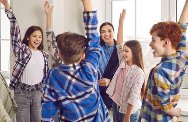 Group of happy junior school students raising hands together while standing in classroom. Cheerful smiling boys, girls classmates and theor teacher having fun together at school during break