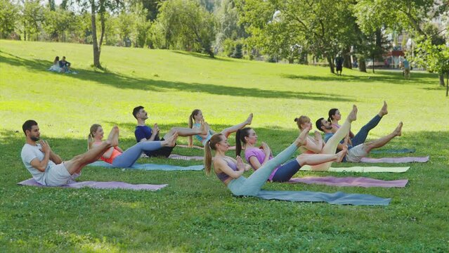 Group of young sporty people practicing yoga lesson with instructor, stretching in Paripurna Navasana exercise. Diverse group sitting and balancing in Boat pose in city park. Concept healthy lifestyle