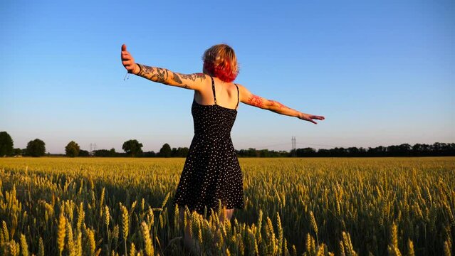 Young hippie woman in dress raising hands while goes among wheat meadow and enjoys freedom. Carefree punk girl with tattoos walking through green barley field at sunset. Beautiful view of countryside
