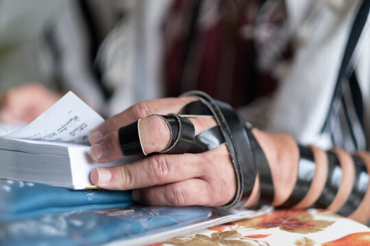 A Jewish man does his morning prayer, with a closeup on his tefillin wrapped around his hand. He is reading through the pages of a siddur, a Jewish prayer book, in the natural light of Jerusalem.