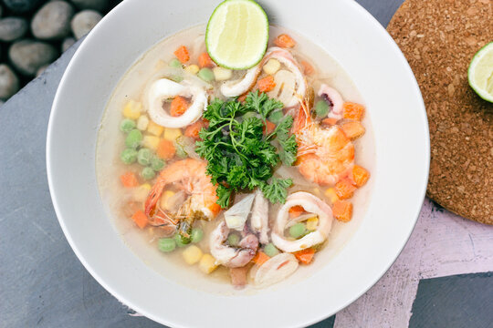 seafood soup in white bowls, soup containing pieces of ring squid, prawns, peas, carrots topped with green celery leaves and lemon slices in it, served on a gray floor.