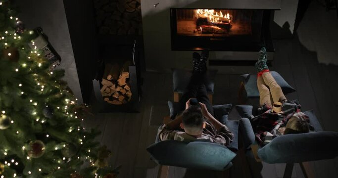A young couple is drinking tea by the fireplace next to a beautiful Christmas tree.
