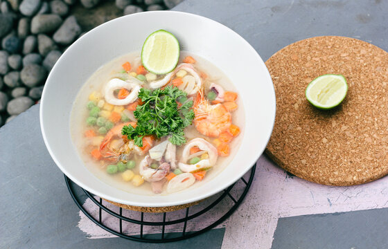 seafood soup in white bowls, soup containing pieces of ring squid, prawns, peas, carrots topped with green celery leaves and lemon slices in it, served on a gray floor.