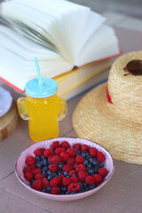 Straw hat, orange soda in the cactus shaped cup, bowl of fresh blueberreis and raspberries, plate of chocolate chip cookies, stack of books and sunglasses in the garden. Selective focus.