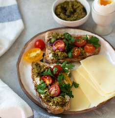 Sandwiches on a plate, bread with pesto, sprinkled with parsley and multi-colored cherry tomatoes, slices of cheese, a soft-boiled egg in a stand, a bowl of pesto, close-up, horizontal.