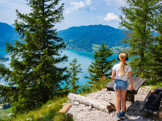 A teenage girl in headphones stands near a bench on a mountain with Lake Attersee in the background, Austria.
