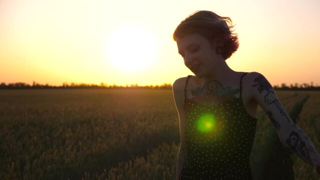 Cheerful punk girl in short black dress fooling around while walks among wheat meadow. Happy female hipster with tattoos having fun during goes through green barley field at sunset. Freedom concept