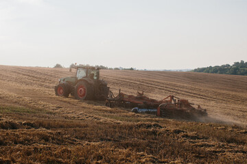 Obraz premium A modern tractor with a heavy trailed disc harrow plows a wheat field at sunset.