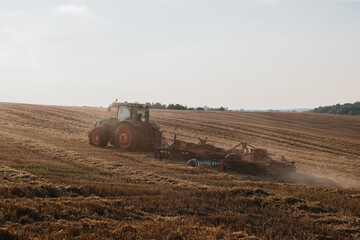 Obraz premium A modern tractor with a heavy trailed disc harrow plows a wheat field at sunset.