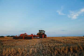 Obraz premium Modern tractor with a heavy trailed disc harrow works a wide hilly field. Autumn or spring agricultural campaign