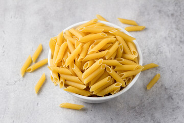 Dry penne pasta in a white ceramic bowl on a gray textured background, top view